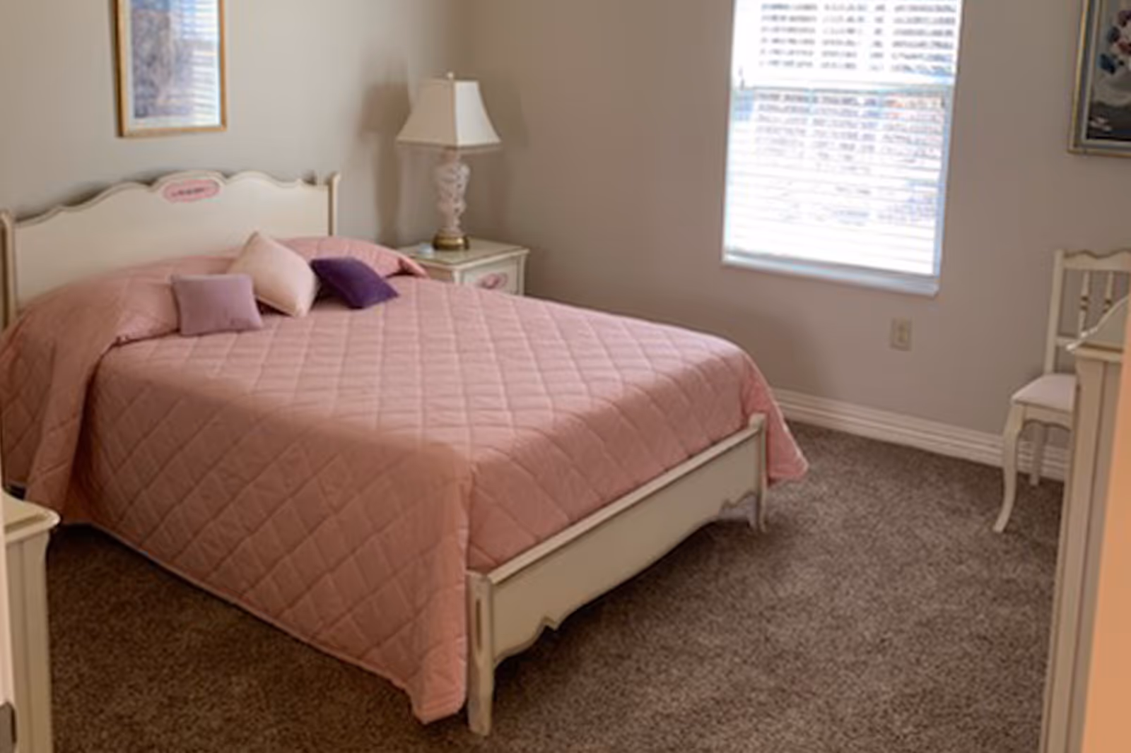 A tidy bedroom with a bed covered in a pink quilt, nightstand with a lamp, and a window with blinds.