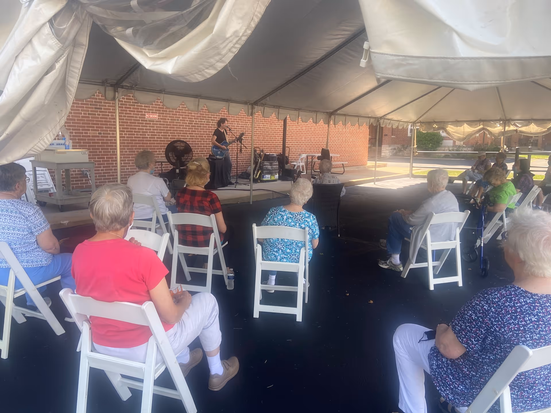 Seniors seated on folding chairs under a large tent watching a musician perform on a small stage.
