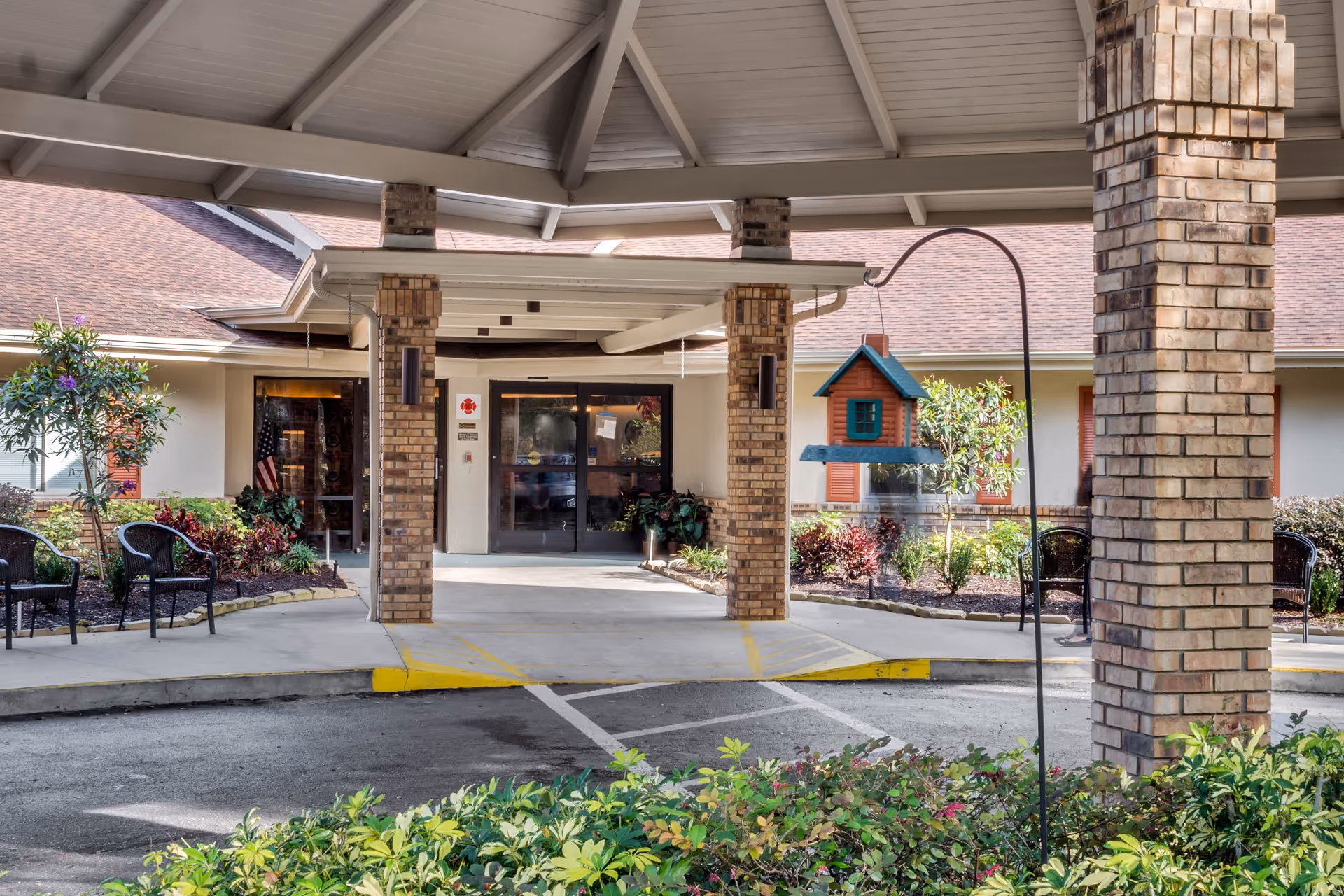 Entrance of Elevated Estates Forest Oaks facility showing a covered drop-off area with brick pillars, a small birdhouse hanging on a metal pole, some outdoor chairs, and landscaped plants and bushes.