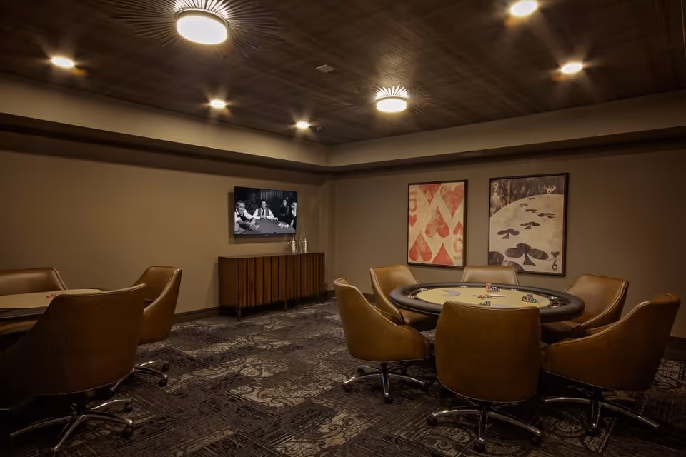 A dimly lit room with a poker table surrounded by six brown leather swivel chairs. On the wall behind the table are two large framed playing card artworks. A flat-screen TV mounted on the adjacent wall displays a black and white image of people playing cards. Below the TV is a wooden cabinet with glassware on top. The ceiling has recessed lighting and two circular ceiling lights.