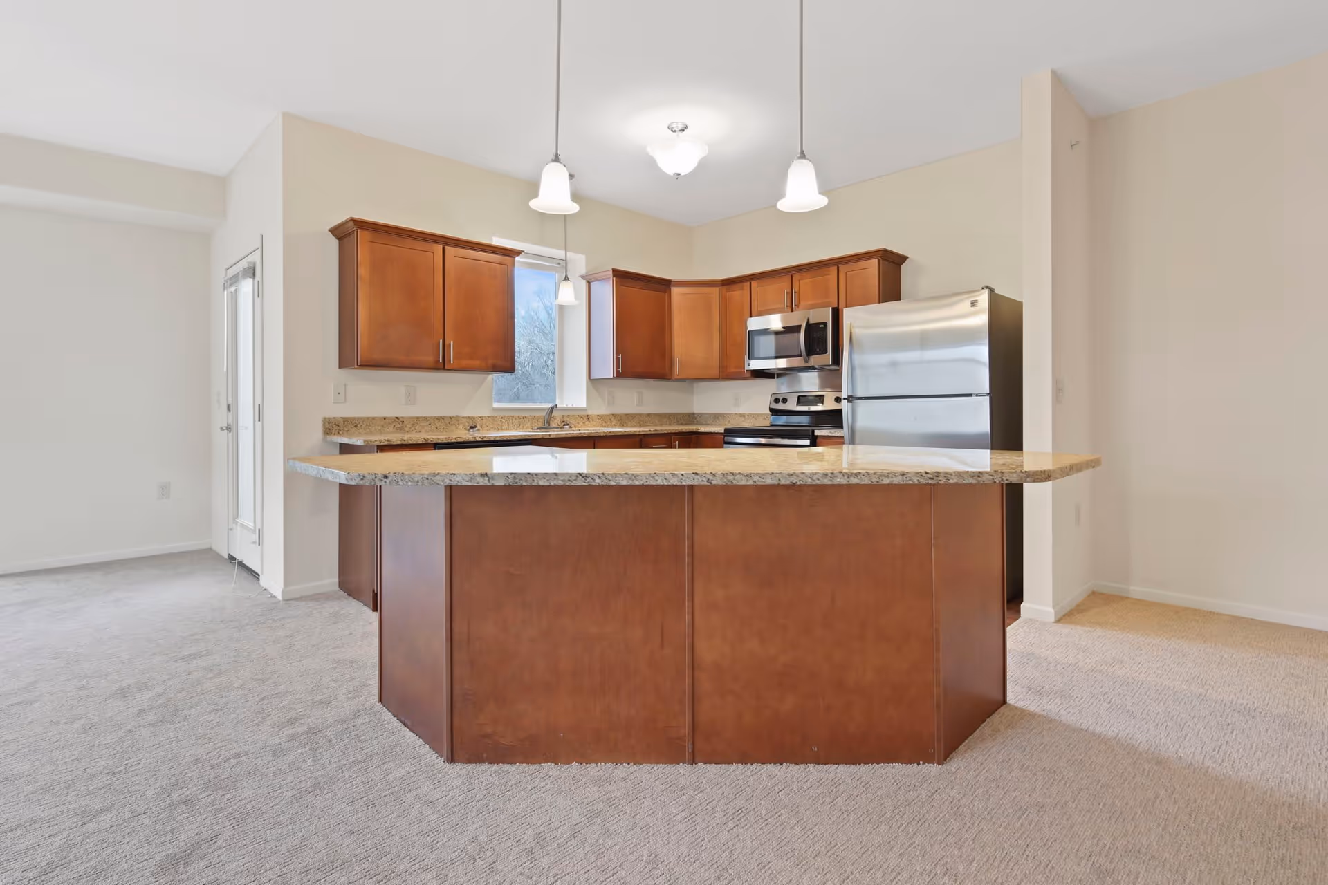 A modern kitchen with wooden cabinets, granite countertops, and stainless steel appliances including a refrigerator, microwave, and stove. The kitchen features a large island with a granite countertop and pendant lighting above. The floor is carpeted and there is a door and window providing natural light.