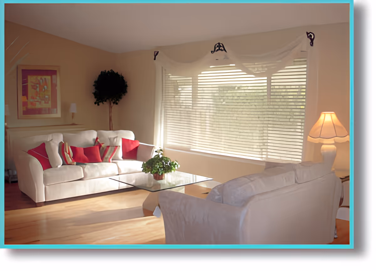 A cozy living room with two beige sofas adorned with red and striped cushions, a glass coffee table with a potted plant, a floor lamp with a beige shade, a framed abstract painting on the wall, a tall indoor plant, and a large window with white blinds and sheer curtains allowing natural light to fill the room.
