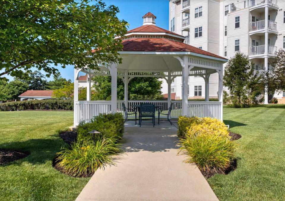 A white wooden gazebo with a table and chairs sits on a paved path amid manicured lawns and landscaping in front of a multi-story senior living building.