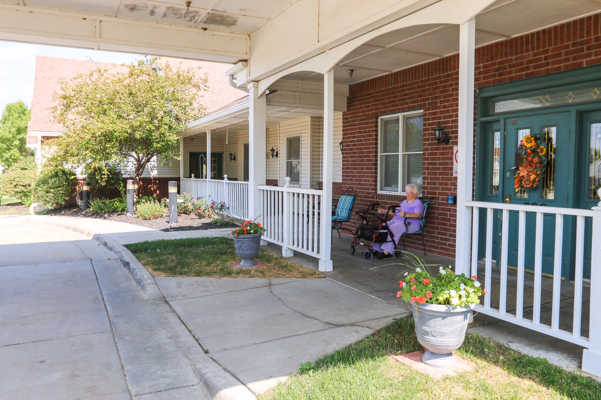 Covered front porch entrance of a senior living facility with potted flowers and an elderly woman seated by a green door.