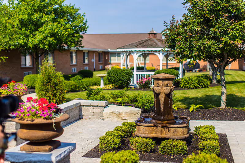 Outdoor garden area with a stone fountain surrounded by neatly trimmed bushes and flower pots with pink and red flowers. There is a white gazebo in the background and a brick building behind it. The scene is sunny with green trees and well-maintained landscaping.