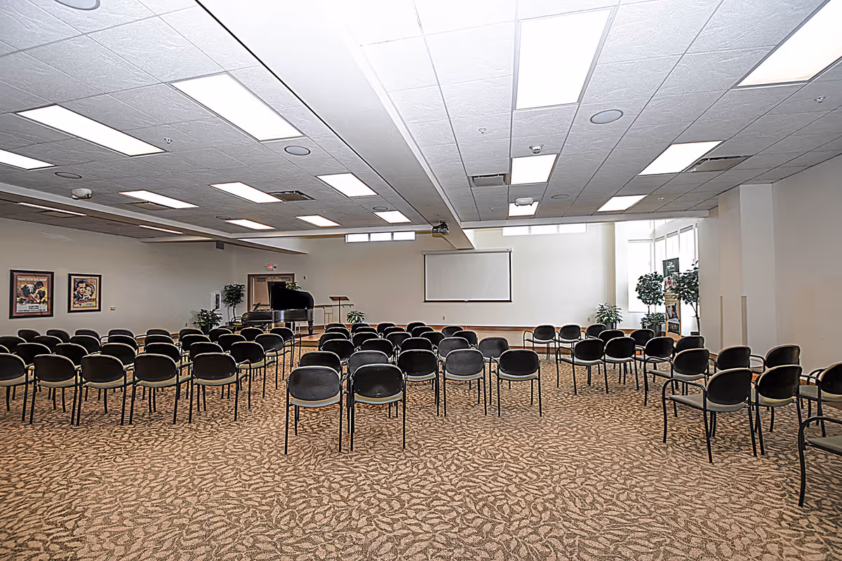 A large meeting or event room with rows of black chairs arranged facing a blank projector screen on the far wall. The room has a patterned carpet, white walls, and a ceiling with multiple rectangular fluorescent lights. There is a grand piano and a music stand near the screen, along with some potted plants and framed pictures on the walls.
