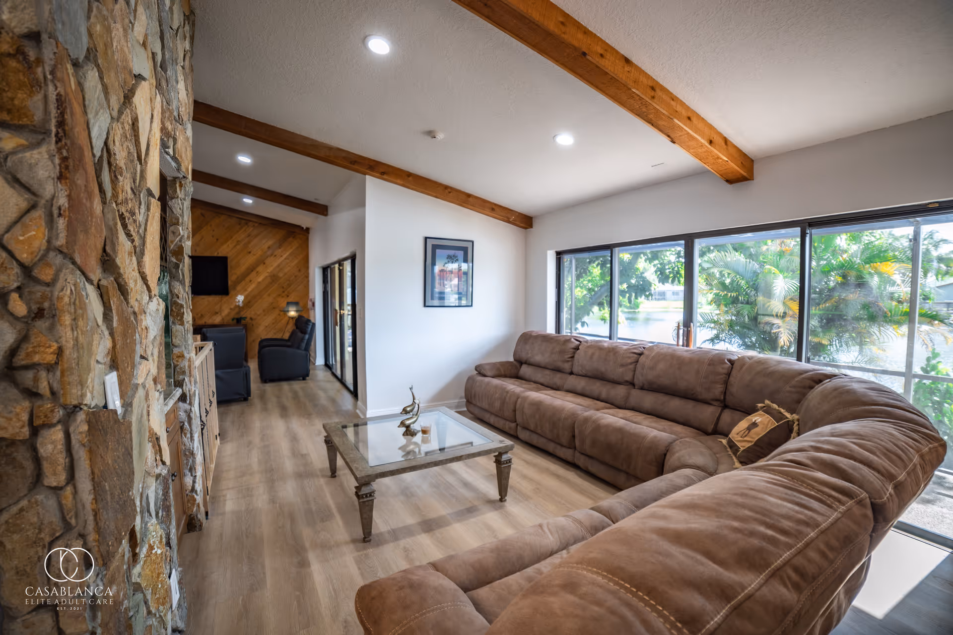 A spacious living room with a large brown sectional sofa and a glass coffee table in the center. The room features wooden ceiling beams, a stone wall on the left, and large windows on the right that provide a view of greenery outside. The floor is covered with light wood flooring, and there is a framed picture on the white wall. In the background, there are two black recliner chairs and a TV mounted on a wooden accent wall.