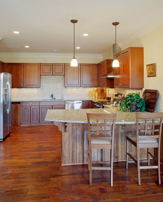 A spacious kitchen with wooden cabinets, a granite countertop island with two wooden chairs, stainless steel refrigerator and dishwasher, pendant lights hanging from the ceiling, and decorative items including plants and vases.