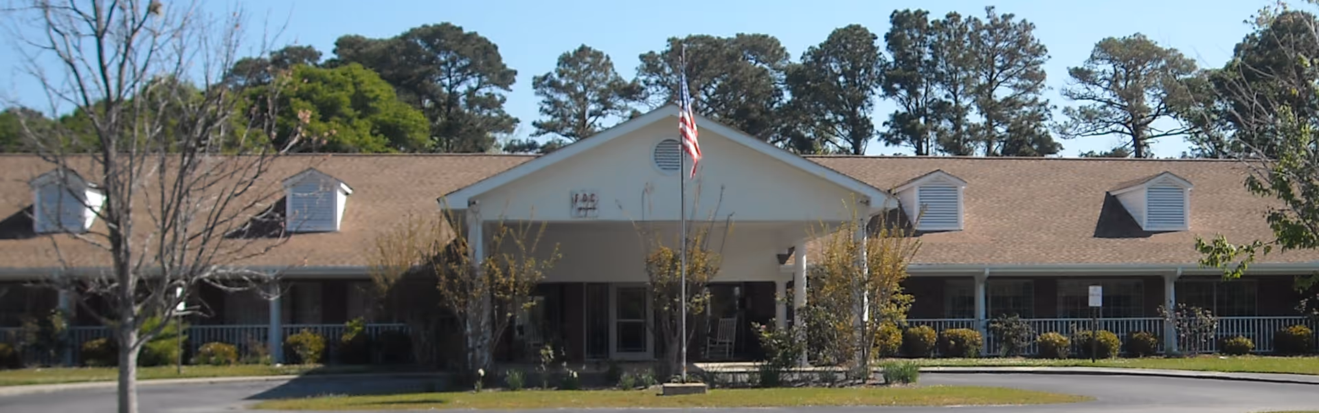 Front exterior view of a single-story building with a peaked roof and dormer windows, surrounded by trees and bushes, with an American flag flying on a flagpole in front.