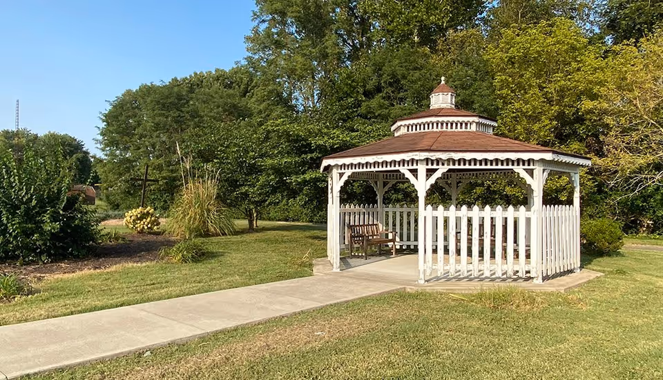 A white wooden gazebo with a brown roof situated on a concrete path in a grassy area surrounded by trees and bushes under a clear blue sky.