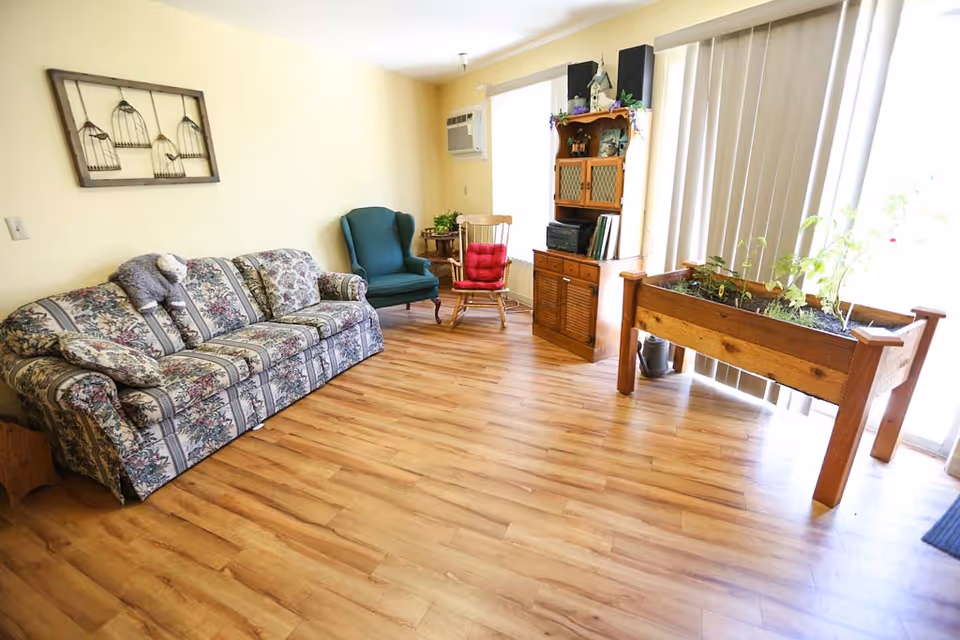 A cozy living room with a floral patterned sofa, a green armchair, and a wooden rocking chair with a red cushion. There is a wooden cabinet with decorative items and books, a wooden planter box with plants near vertical blinds covering a window, and a wall-mounted air conditioning unit. The floor is light wood, and the walls are painted a soft yellow.