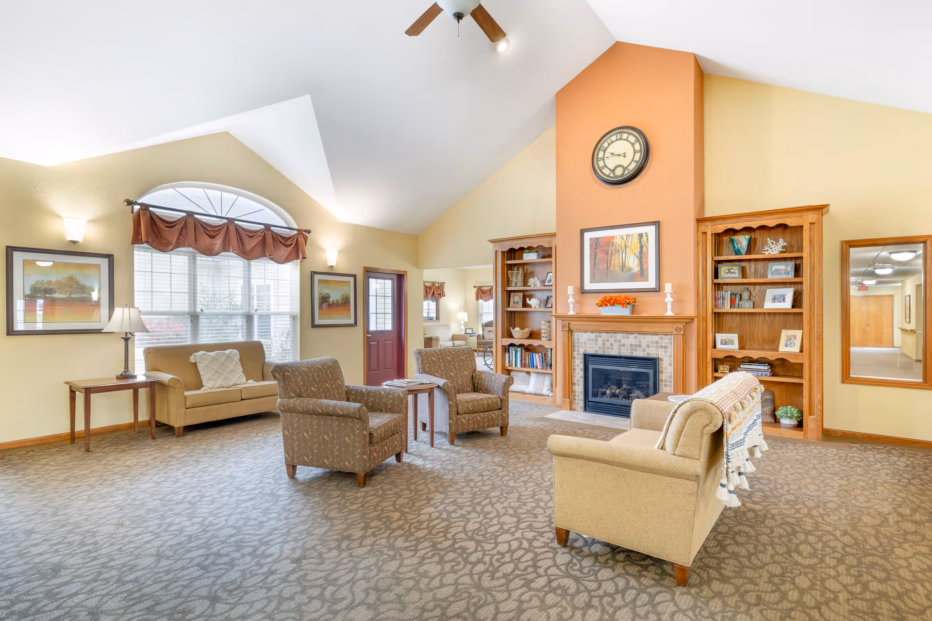 A bright and spacious living room area in an assisted living facility featuring a high vaulted ceiling with a ceiling fan, a large arched window with brown curtains, beige walls, and carpeted floor. The room is furnished with two beige sofas, two patterned armchairs, a wooden coffee table, and side tables with lamps. A fireplace with a tiled surround is centered between two wooden bookshelves filled with books and decorative items. Above the fireplace is a framed painting and a round wall clock. A mirror hangs on the wall to the right, reflecting a hallway.