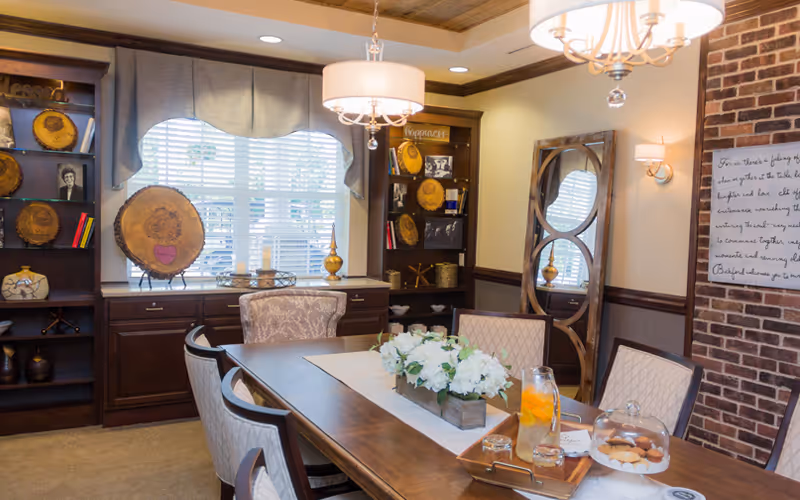 A warmly lit dining room with a wooden table set with a white runner, a centerpiece of white flowers, a pitcher of iced tea with lemon slices, and a tray of cookies under a glass dome. The room features upholstered chairs around the table, built-in dark wood shelves with decorative items and framed photos, a large window with blinds and a valance, a tall decorative mirror, and a brick accent wall with a framed inspirational quote. Two elegant chandeliers hang from the ceiling.