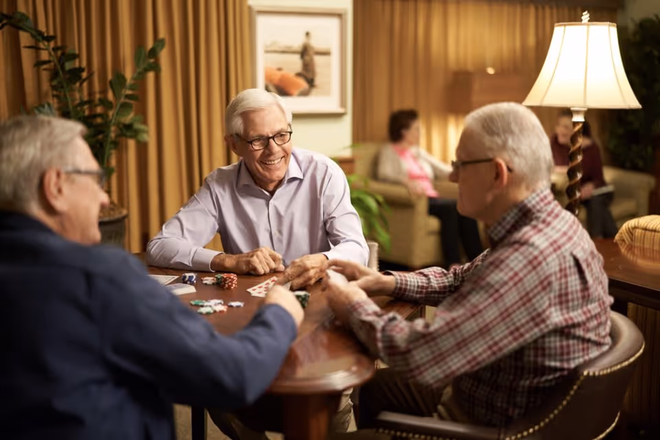 Three elderly men sitting around a wooden table playing cards and poker chips in a warmly lit living room with beige curtains, a lamp, and two women sitting on a couch in the background.
