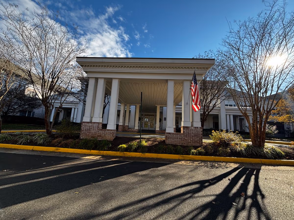 Front entrance porte-cochere of a senior living community with white columns, an American flag, and trees under a blue sky.