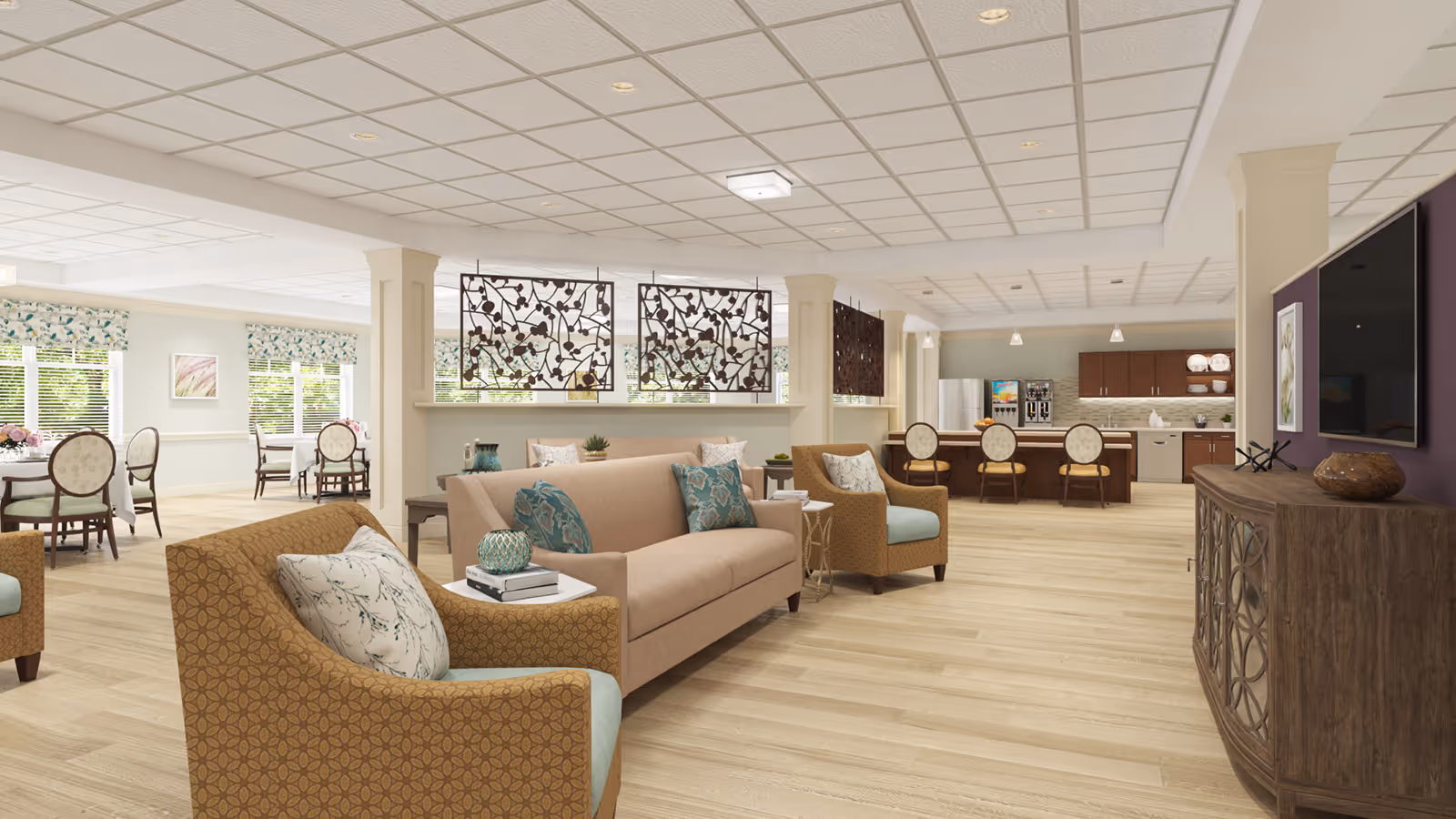 A spacious and well-lit common area in a senior living facility featuring comfortable seating with patterned armchairs and a beige sofa adorned with decorative pillows. The room includes a kitchen area with a refrigerator, coffee machines, and cabinetry. Decorative metal panels hang above a half wall separating the seating area from the dining area, which has tables and chairs near windows with floral valances. A large flat-screen TV is mounted on a purple accent wall above a wooden cabinet.