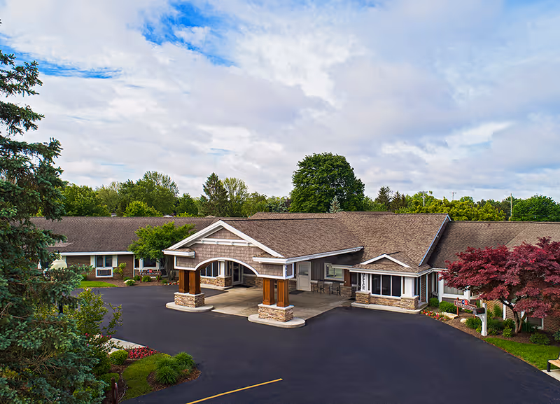 Front entrance of a single-story senior living facility with a covered porte-cochere, landscaped grounds, and surrounding trees.