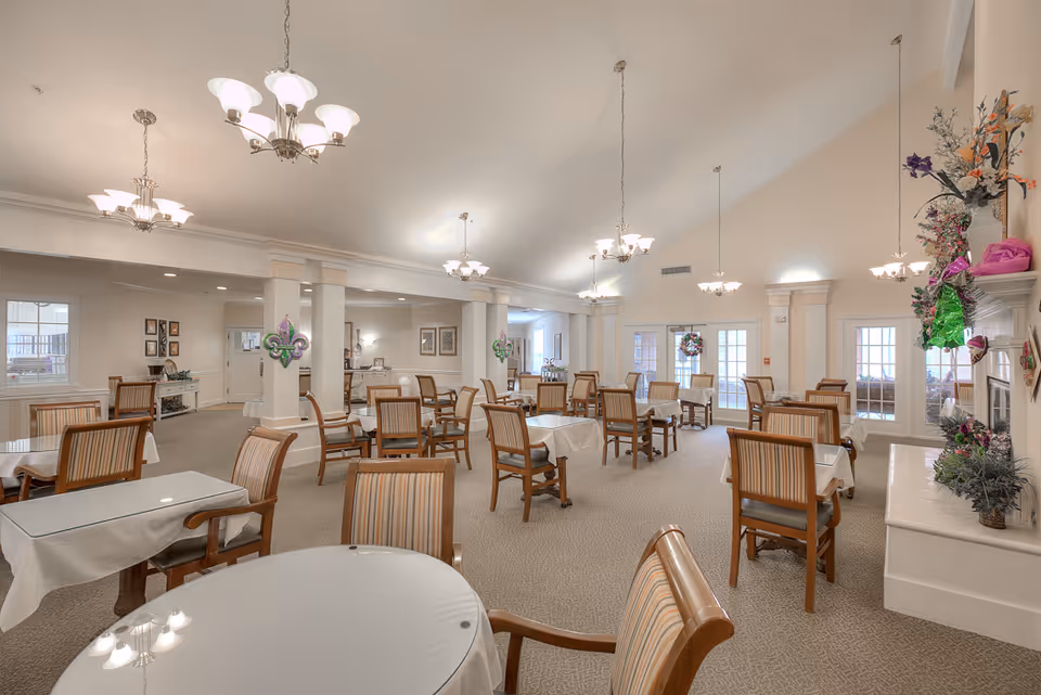 A spacious dining room with multiple tables covered with white tablecloths and surrounded by wooden chairs with striped cushions. The room has beige walls, carpeted floors, and several chandeliers hanging from a high ceiling. There are decorative columns, framed pictures on the walls, and a fireplace adorned with flowers and decorations. Large windows and glass doors allow natural light to enter the room.