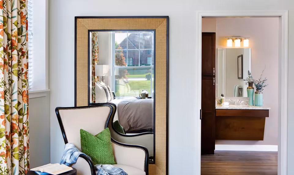 Cozy seating area with an armchair and decorative mirror reflecting a bed, with a bathroom visible through an open door.