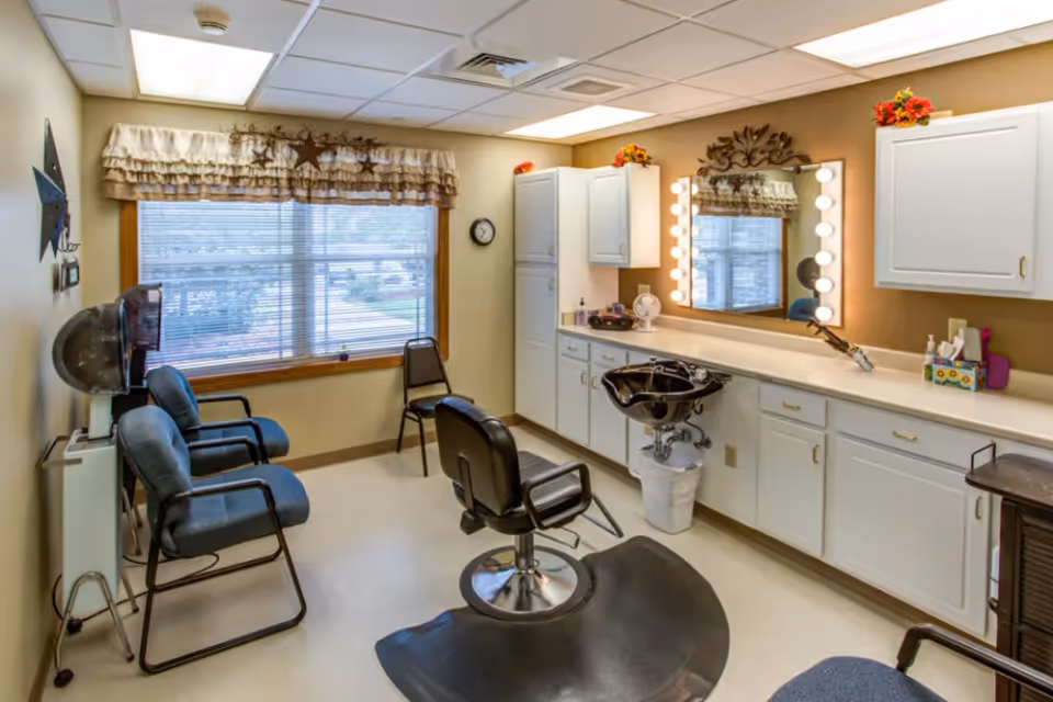 Small salon room with a styling chair and shampoo sink in front of a lighted vanity mirror, cabinets, and waiting chairs by a large window.