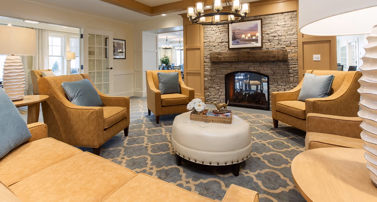 A cozy communal living room with tan leather armchairs arranged around a central ottoman in front of a stone fireplace.
