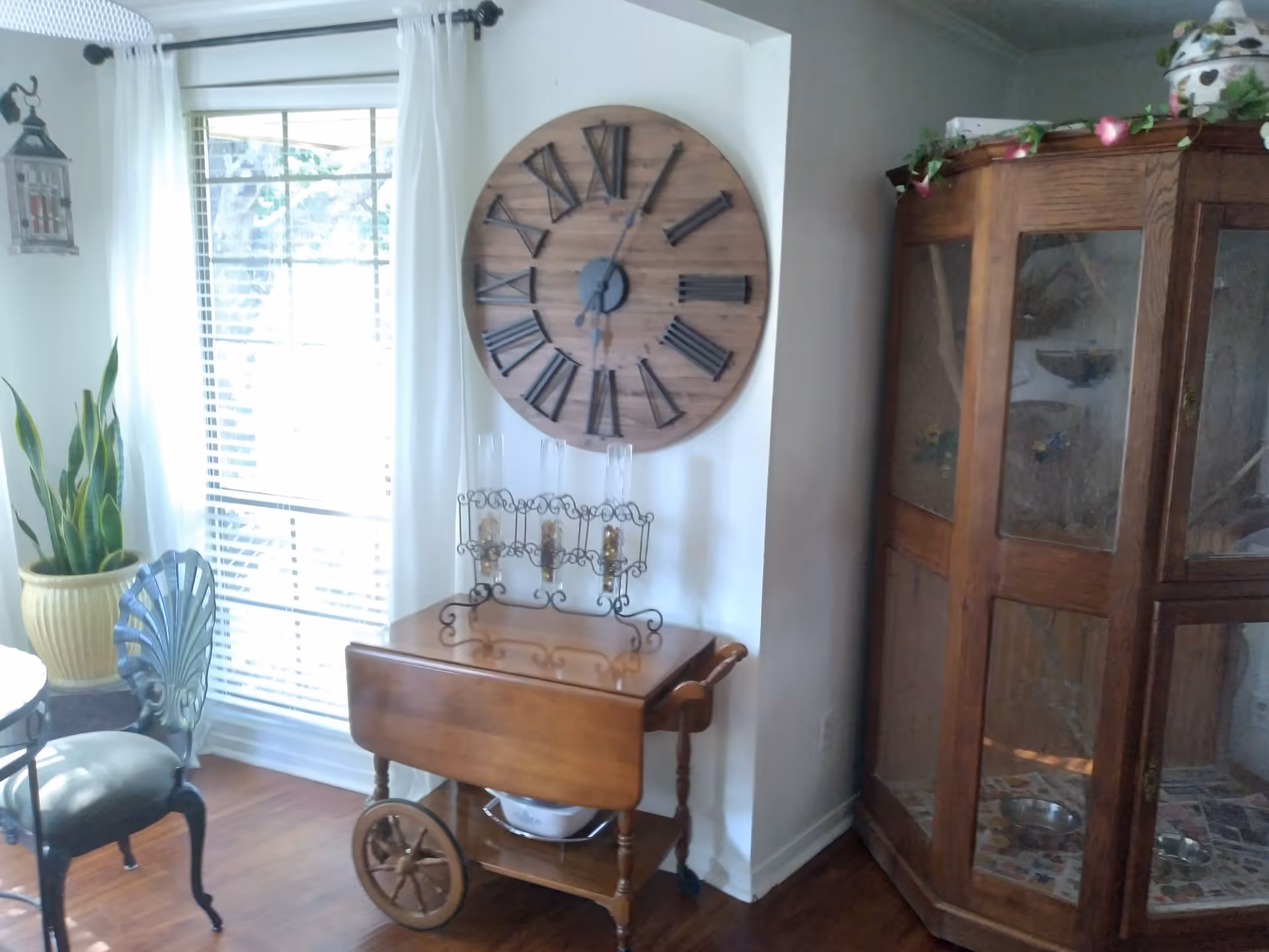 Sunlit dining corner showing a large round wooden wall clock above a wooden serving cart, glass-front cabinet, dining chairs, and a potted plant by a window.