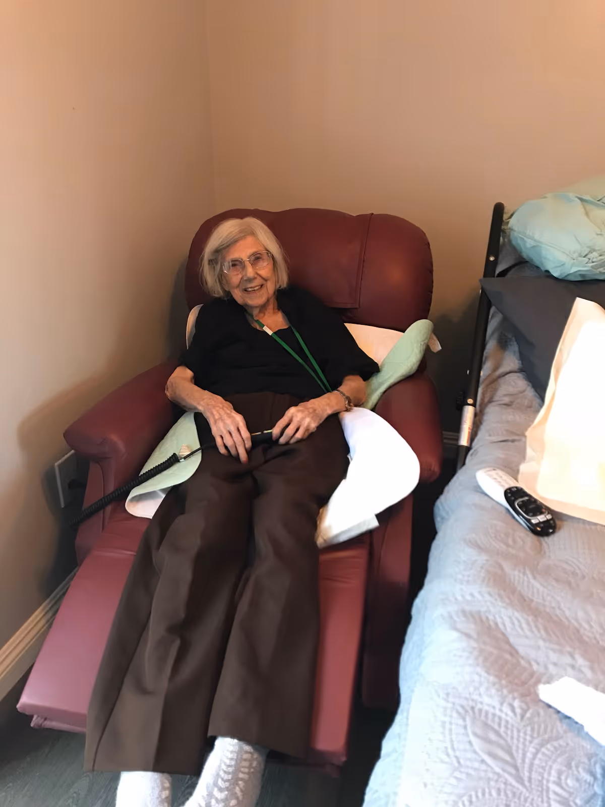 An elderly woman with gray hair and glasses is reclining in a maroon armchair next to a bed with a gray quilt. She is smiling and holding a remote control device, with pillows supporting her back and sides.