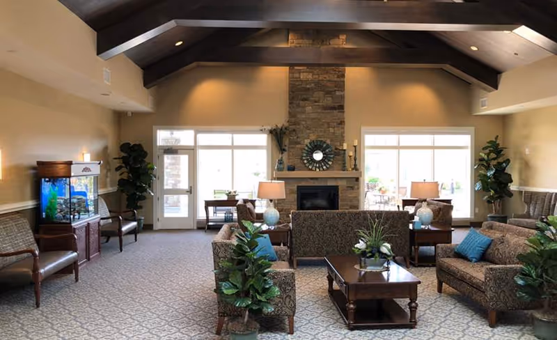 Spacious furnished common living room with multiple seating areas, a stone fireplace, vaulted beamed ceiling, plants, and an aquarium.