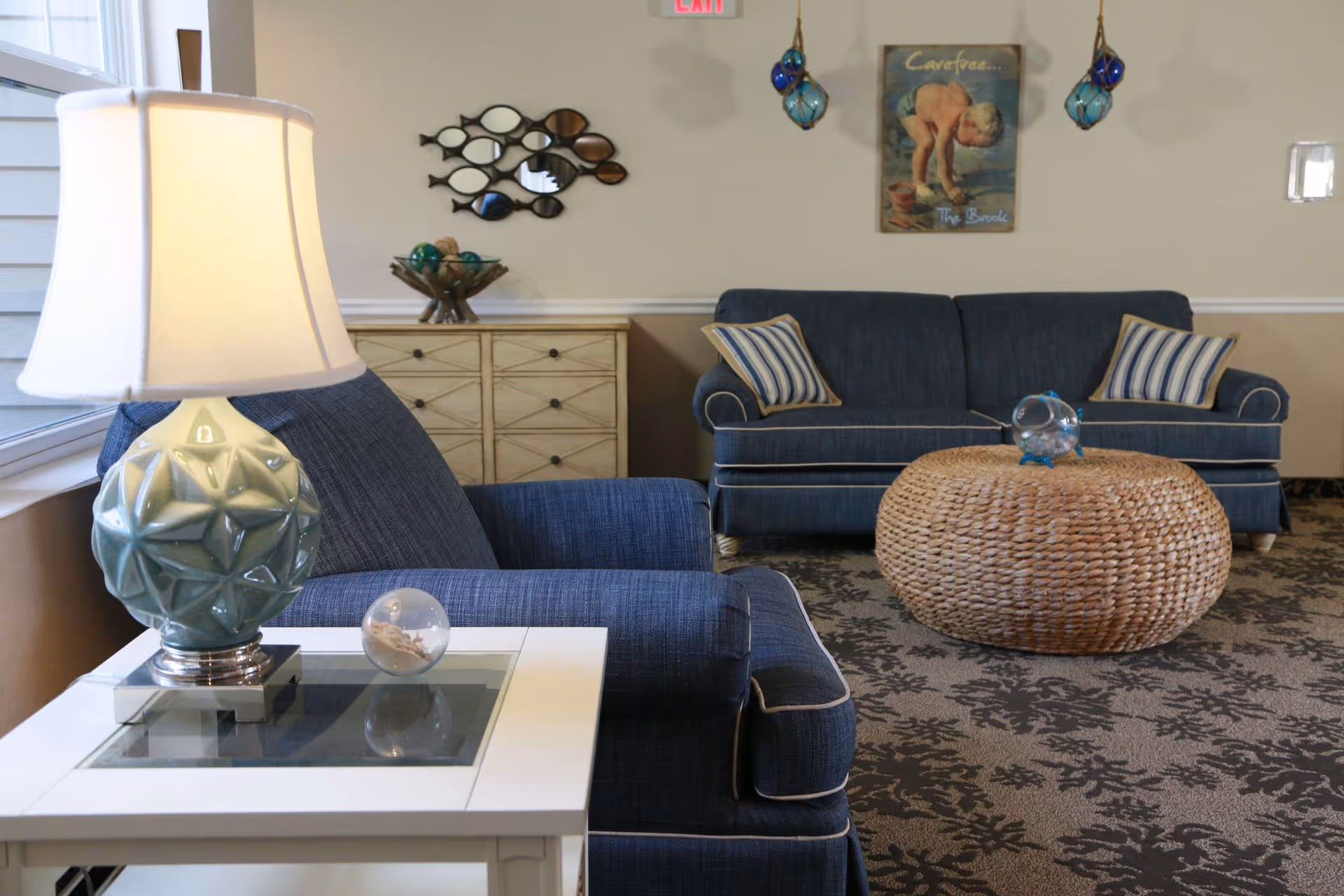 A cozy living room area with blue upholstered armchairs and a matching sofa adorned with striped pillows. A round woven coffee table sits in front of the sofa, topped with a decorative glass orb. A white side table with a blue ceramic lamp and a small decorative glass sphere is next to the armchair. The room has a patterned carpet, a light-colored wall with a fish-shaped mirror, a wooden cabinet, and a vintage-style poster on the wall. Two blue hanging glass ornaments are also visible.