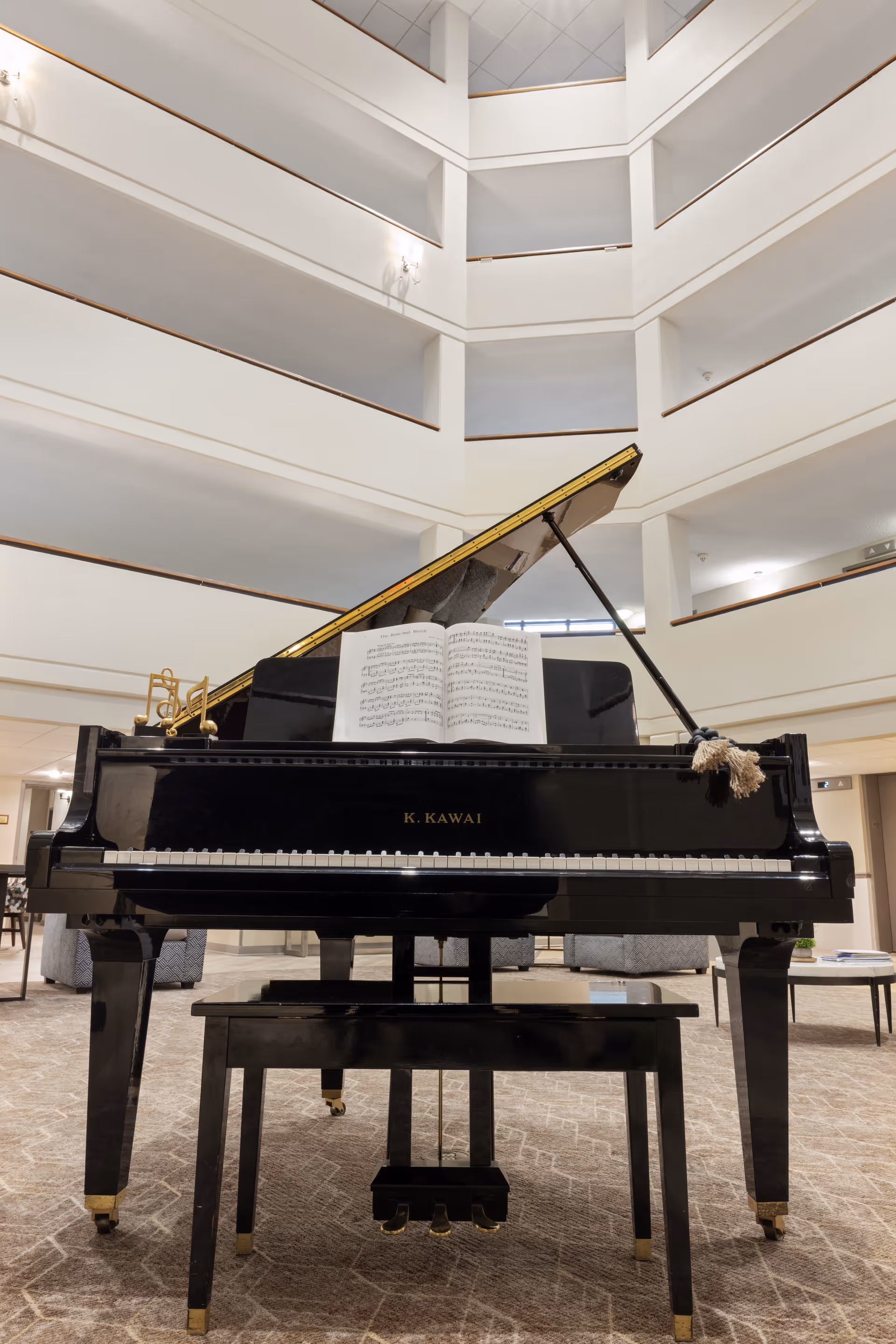 A black K. Kawai grand piano with sheet music sits in the multi‑story atrium/lobby of a senior living facility.