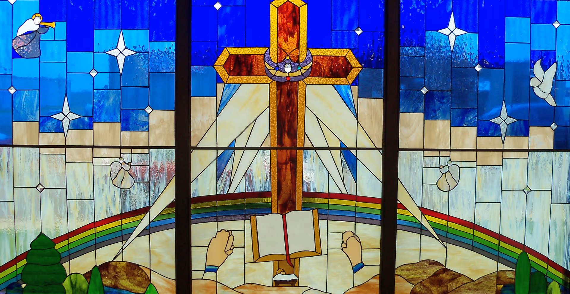 A stained glass window featuring a large cross with a crown of thorns at the center, rays of light shining from behind it. Below the cross is an open book with two raised fists on either side. The background includes a rainbow arching across the lower part, blue sky with stars, angels, and a white dove.