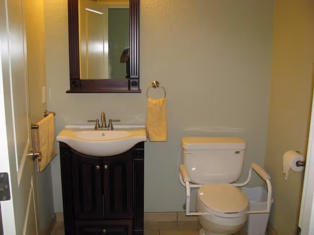A small bathroom with a white toilet equipped with safety grab bars on both sides, a dark wood vanity with a white sink, a mirror above the sink, and yellow hand towels hanging on a ring and a bar.