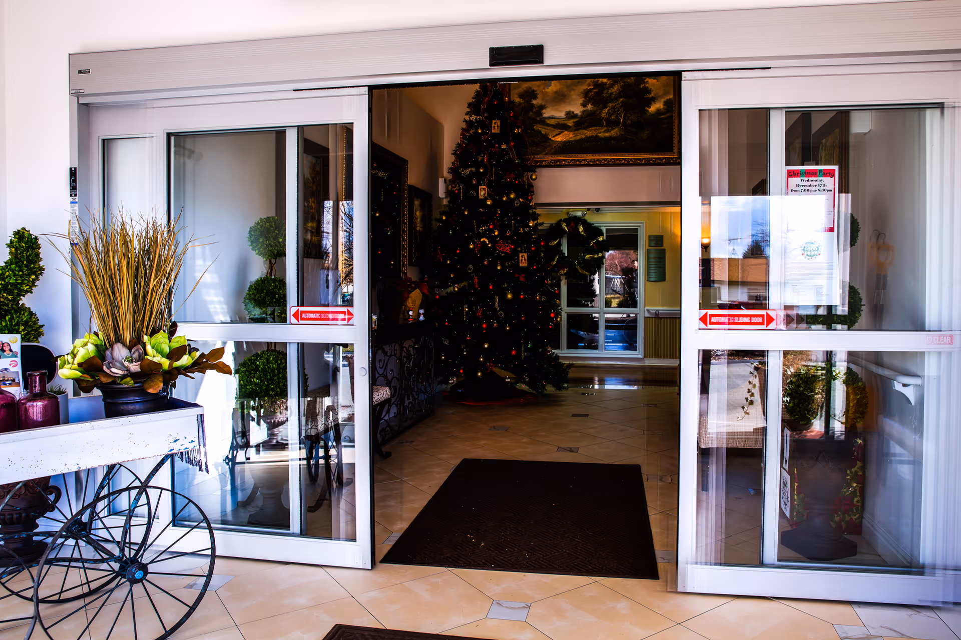 Sliding glass entrance doors opening into a decorated lobby with a large Christmas tree and potted plants.