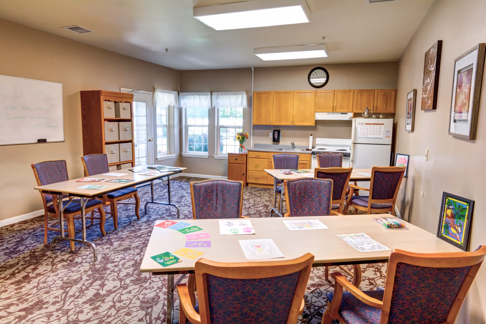 A well-lit room with multiple tables and chairs arranged for group activities. The tables have coloring sheets and art supplies on them. The room features a small kitchenette with wooden cabinets, a refrigerator, and a stove. There are windows with white curtains letting in natural light, a whiteboard on one wall, and framed artwork on another wall.