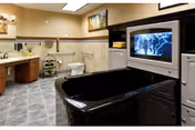 A spacious bathroom featuring a large black bathtub with a built-in television screen at the foot of the tub. The room has tiled flooring and walls with a vanity area including a sink, mirror, and lighting fixtures. There is also a toilet and a small shelving unit with towels and decorative items.