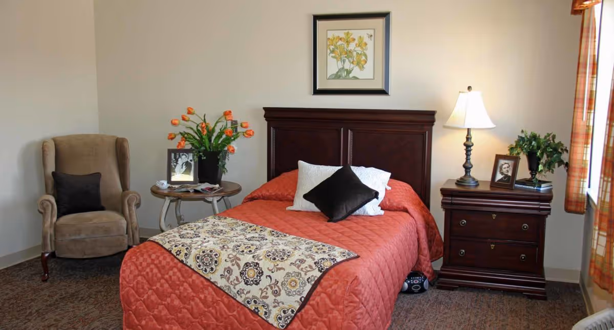 A cozy bedroom with a single bed covered in a red quilt and decorative pillows. There is a wooden headboard, a nightstand with a lamp, framed photo, and a plant. Next to the bed is a small round table with a vase of orange flowers and a framed photo. A beige upholstered armchair with a dark cushion is placed in the corner. The room has beige walls and a window with plaid curtains letting in natural light.