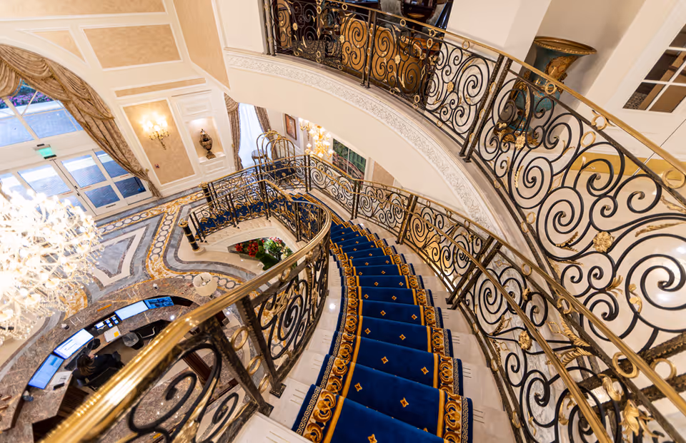 Ornate curved marble staircase with blue carpet and gold-and-black wrought-iron railing overlooking a chandelier-lit lobby and reception desk.