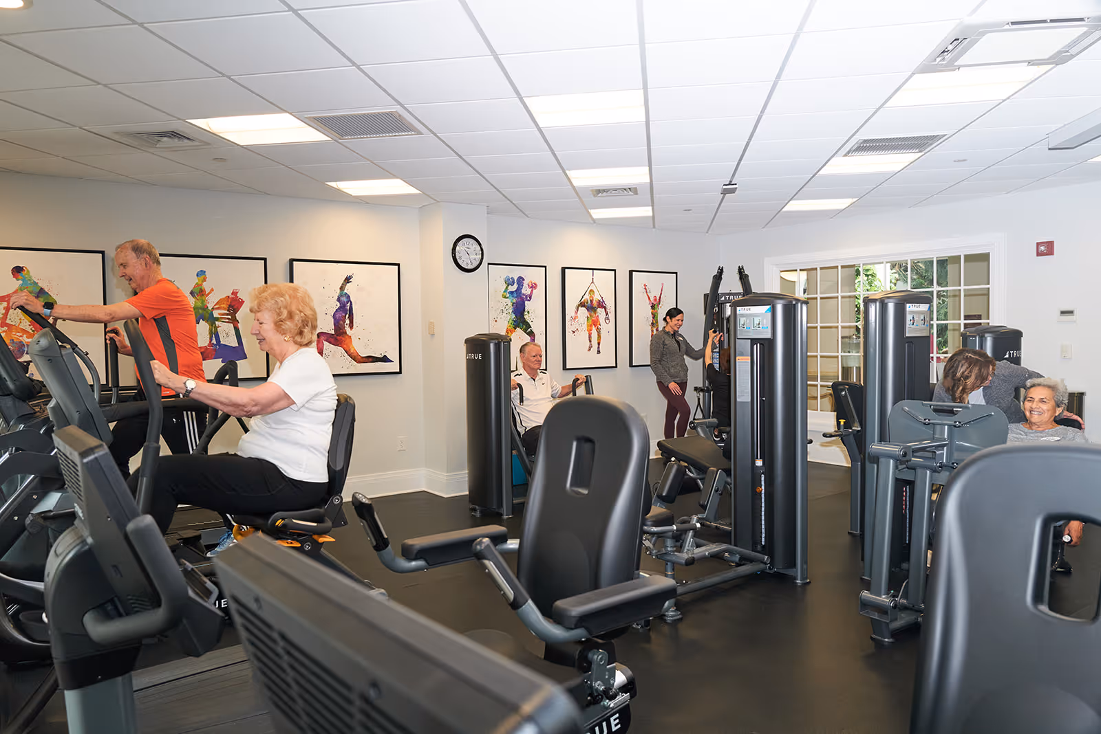 A fitness room in a senior living facility with several elderly people using exercise equipment including stationary bikes and weight machines. The room has white walls decorated with colorful artwork of people in various athletic poses. There is a clock on the wall and large windows letting in natural light.