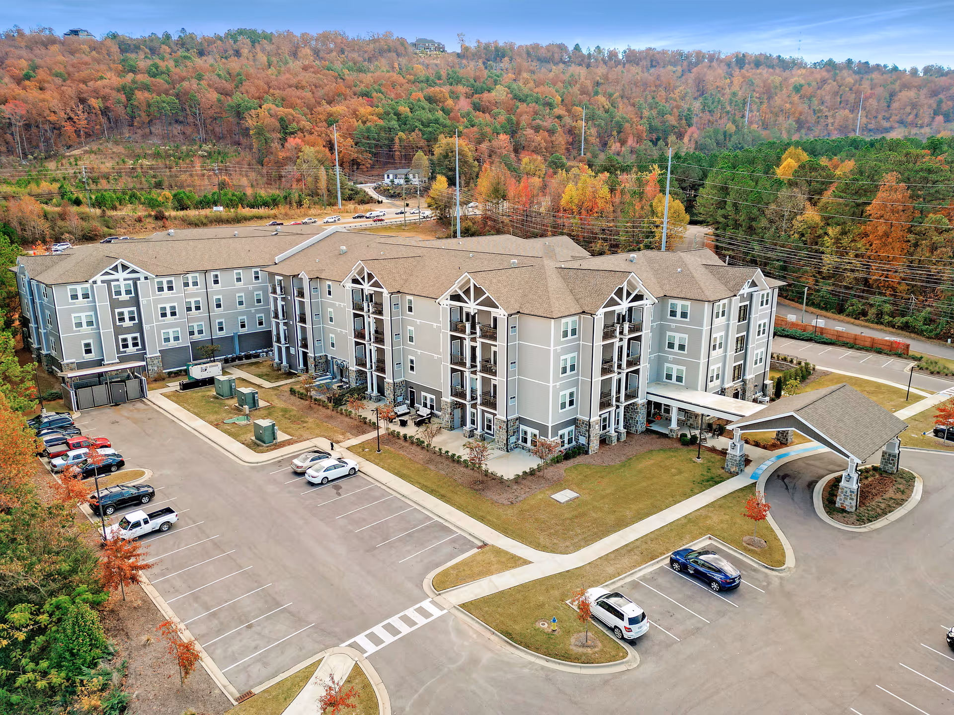 Aerial view of Monark Grove Greystone, a multi-story senior living facility surrounded by a parking lot and autumn-colored trees on a hillside in the background.