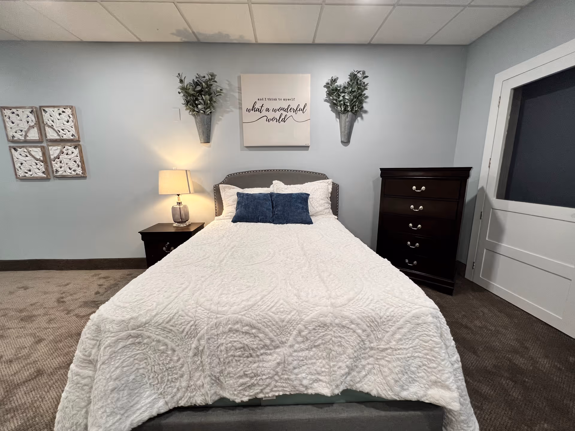 A neatly made bed with a white quilt and blue pillows, bedside table with lamp, wall decor and a dresser in a softly lit bedroom.