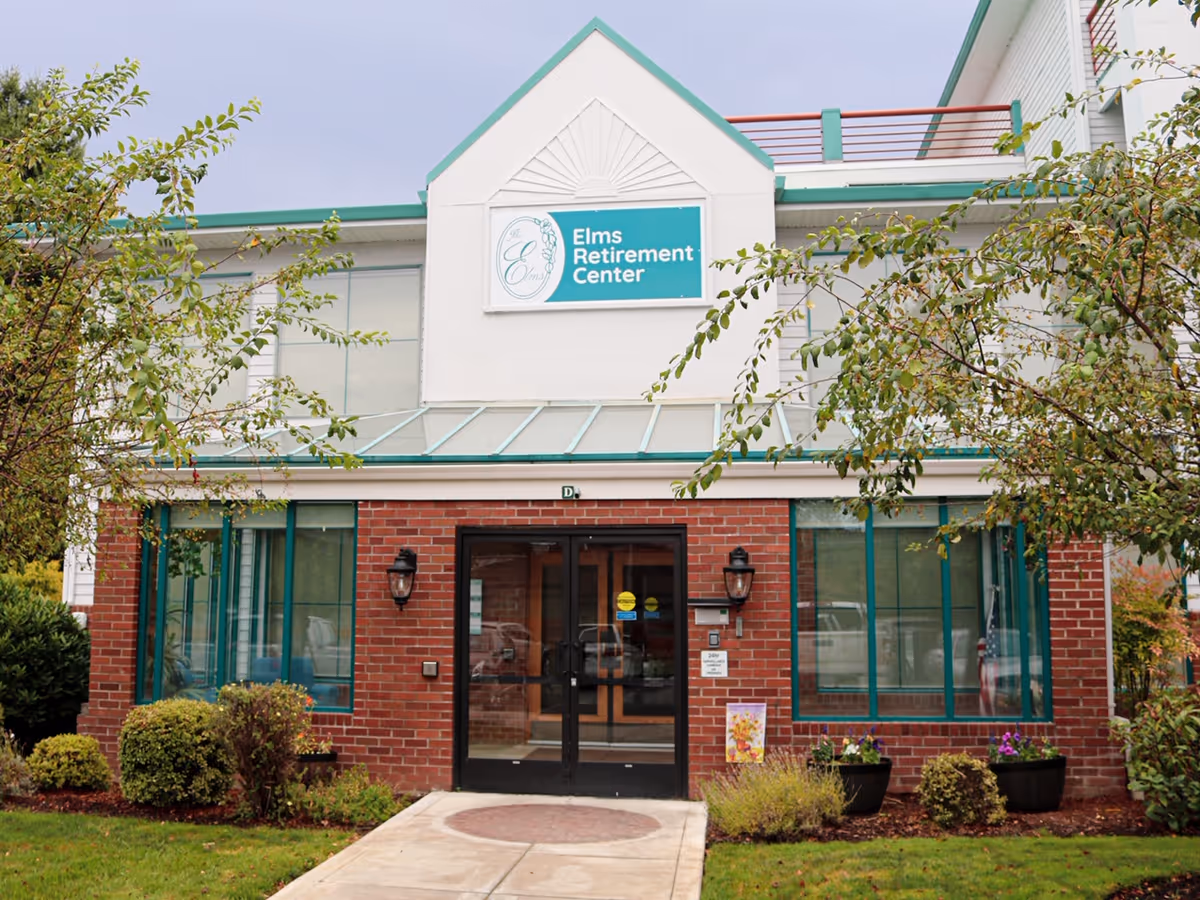 Front entrance of the Elms Retirement Center building with brick facade, glass doors, windows and a sign above.