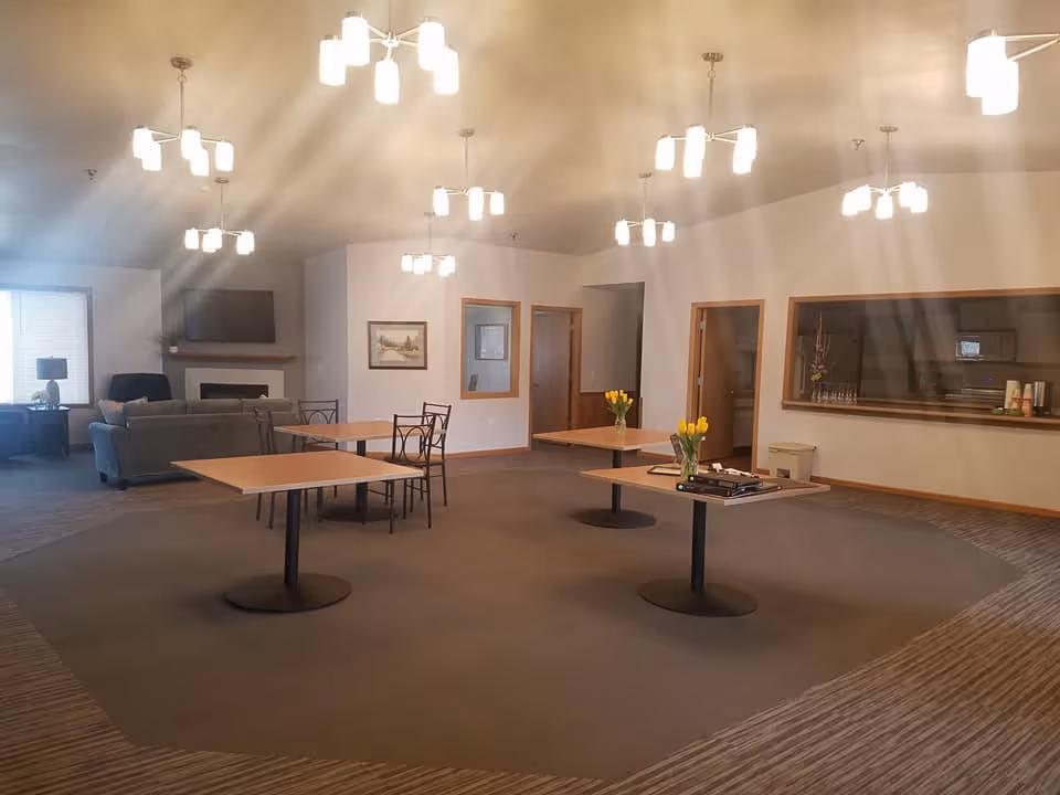 A spacious common area with several square tables, some with yellow tulip flower arrangements, and a few chairs. In the background, there is a seating area with a gray couch, a black armchair, a fireplace, and a wall-mounted TV. The room has multiple ceiling lights and large windows letting in natural light.