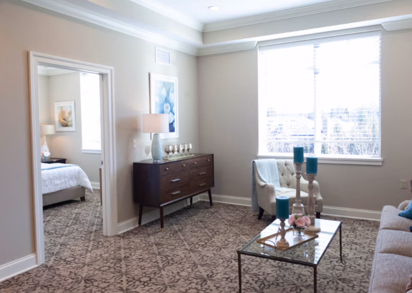 A bright living room with patterned carpet, a glass coffee table with candles and flowers, a beige armchair with a throw blanket, a sofa with blue pillows, and a wooden sideboard with a lamp and decorative items. A doorway leads to a bedroom with a bed and a window.