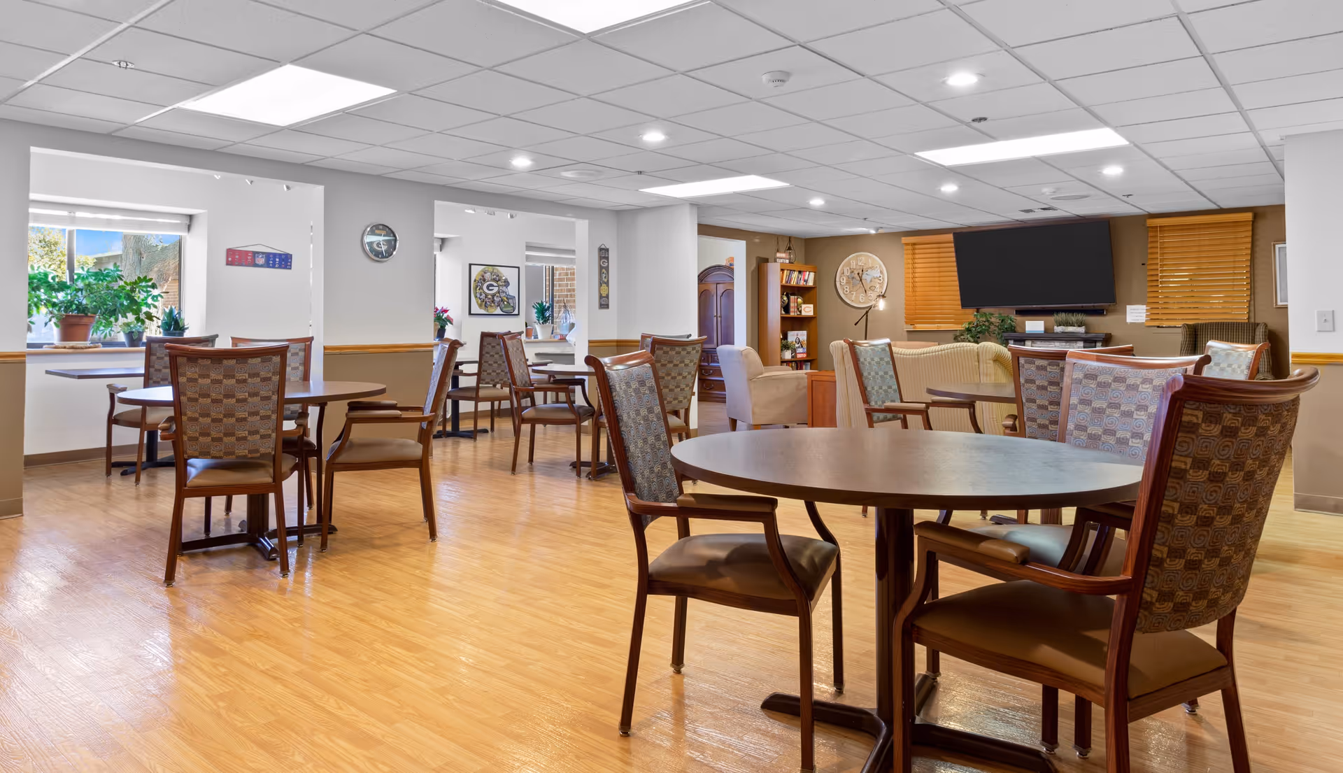 A spacious senior living common area with multiple round wooden tables and cushioned chairs arranged on a wood floor. The room has a drop ceiling with recessed lighting, large windows with plants on the sills, a wall clock, a bookshelf, a large flat-screen TV mounted on a brown accent wall, and comfortable armchairs near the TV.