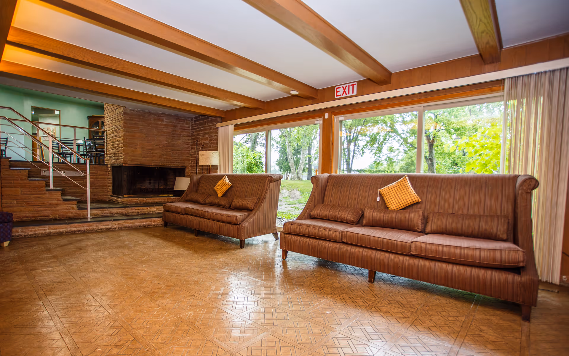 A spacious living room with two brown striped sofas each adorned with a yellow checkered pillow. The room features a large brick fireplace, wooden ceiling beams, and large windows with a view of green trees outside. There is a staircase leading to an upper level with a railing and a lamp beside one of the sofas.