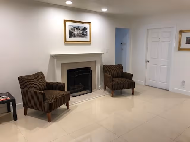 A clean, bright room with two brown striped armchairs placed on either side of a white fireplace with a black screen. Above the fireplace hangs a framed picture. To the right, there is a white door labeled with the number 1 and a small framed picture on the wall. The floor is tiled with large, light-colored tiles, and the walls are painted white.