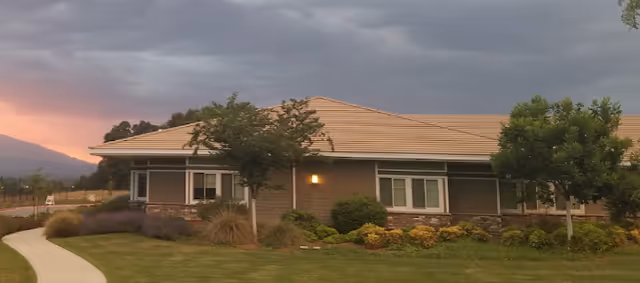 Single-story building with a tan roof and beige walls surrounded by green grass, bushes, and trees under a cloudy sky at sunset.