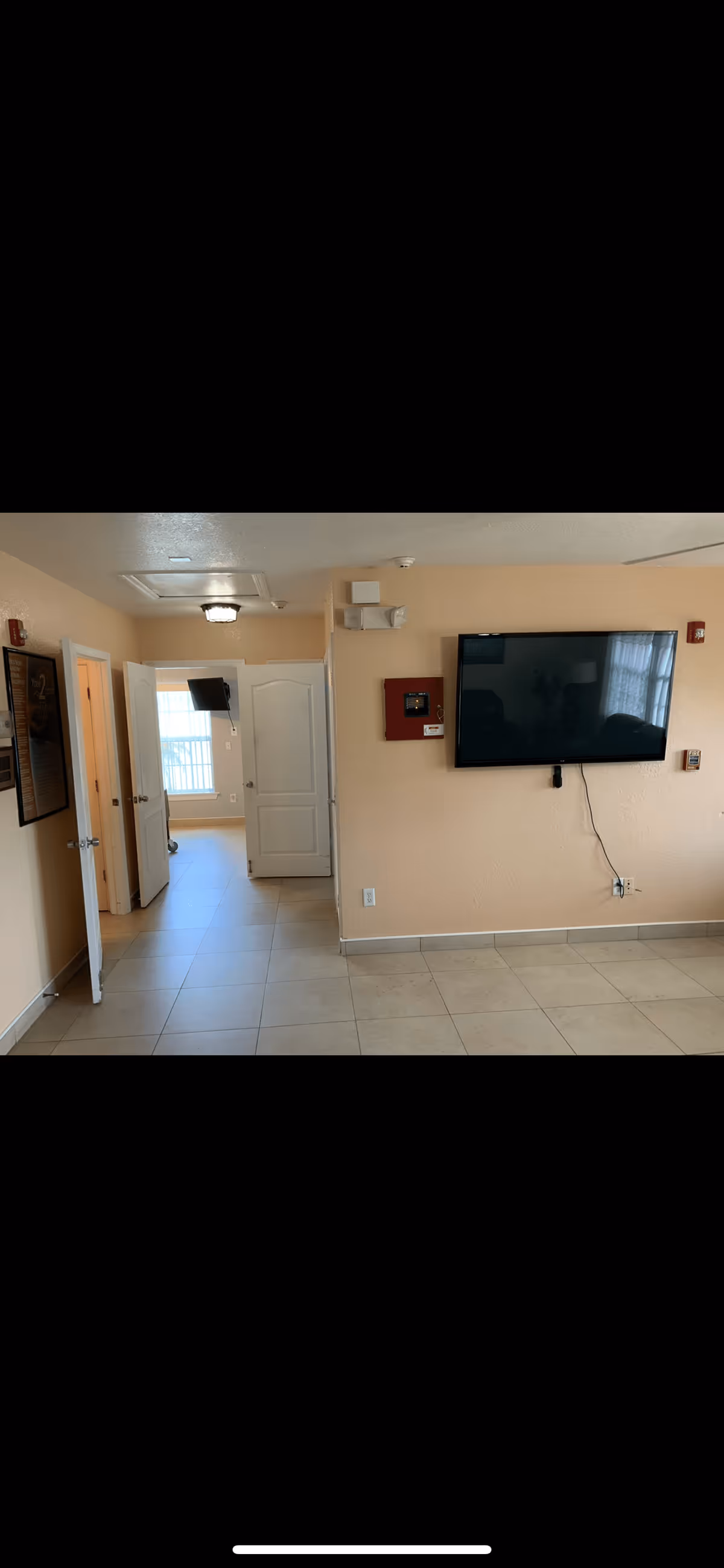 Interior view of a senior living facility hallway with beige walls and tiled floor. There are two open white doors on the left side leading to rooms with windows and a wall-mounted TV. On the right wall, there is a large flat-screen TV mounted above a red fire alarm panel and some electrical outlets. The area is well-lit with natural light coming through the windows.