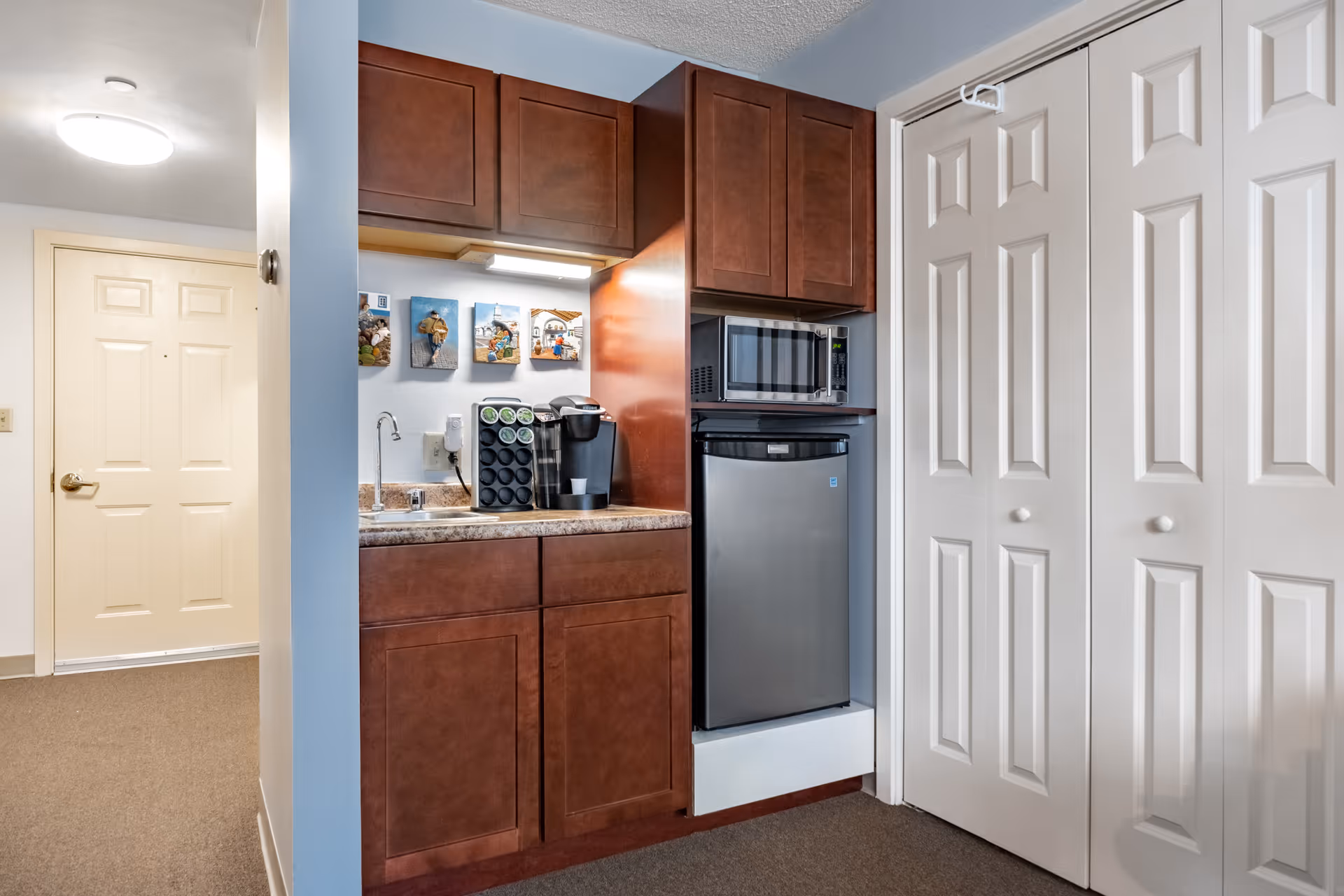 Small kitchenette area with dark wood cabinets, a countertop with a coffee maker and coffee pods, a small sink, a microwave above a compact refrigerator, and white bi-fold closet doors to the right. A beige entry door is visible in the background.