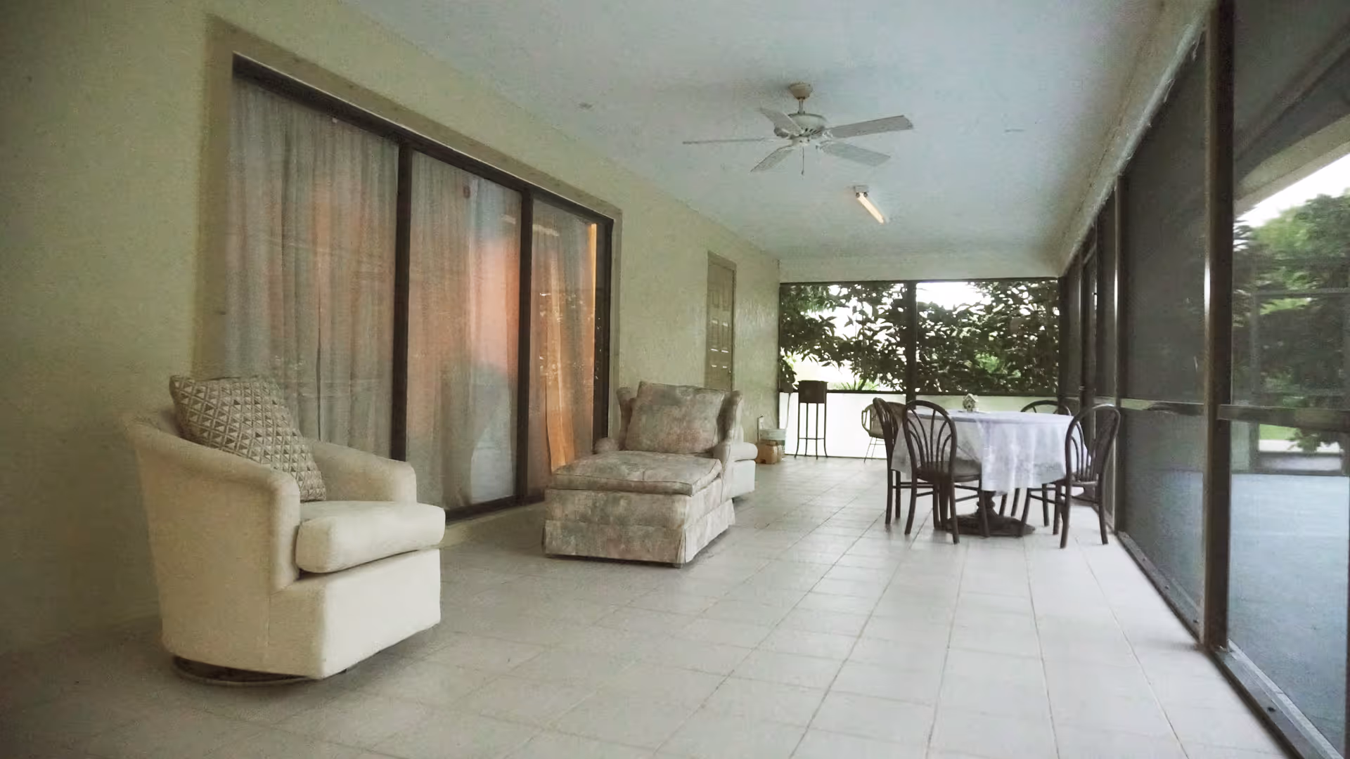 A spacious screened-in porch with tiled flooring, featuring a cushioned armchair with a patterned pillow, a cushioned chaise lounge, and a round table covered with a white tablecloth surrounded by four chairs. Ceiling fan and large sliding glass doors are visible, with greenery outside the screened area.